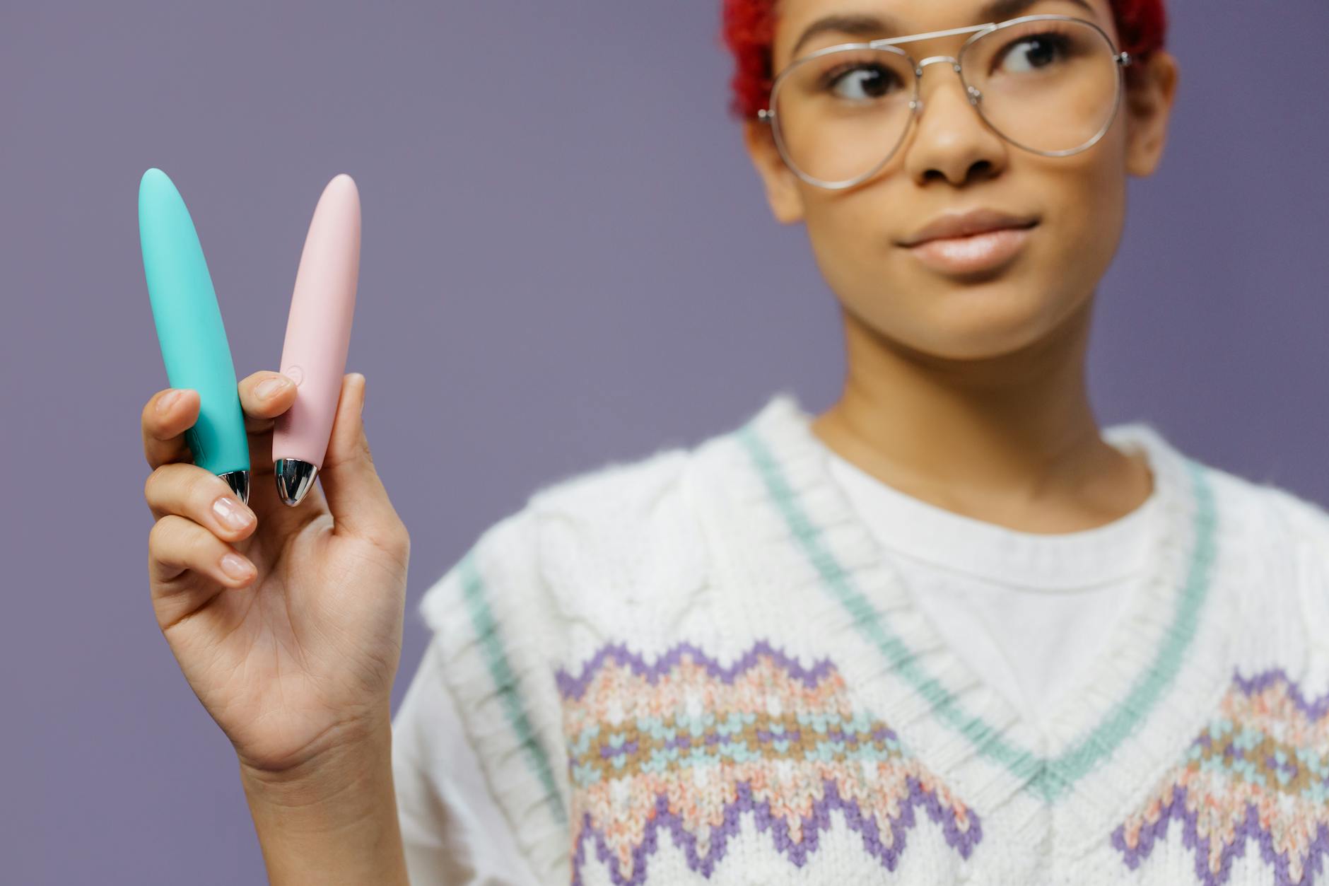 A woman holds two colorful vibrators against a purple background, representing pleasure exploration and sexual wellness